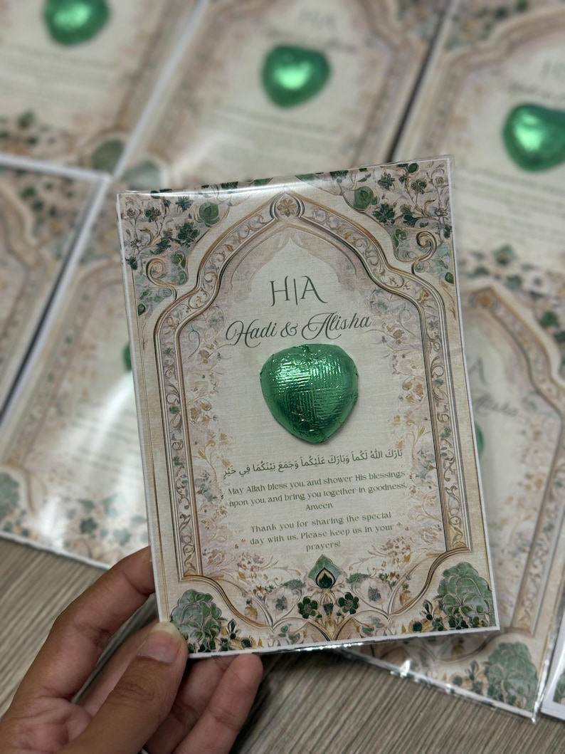 May include: A wedding favor card with a green heart-shaped chocolate. The card has an ornate design with the names "Hadi & Alisha" and text in English and Arabic. The card is held in a hand.