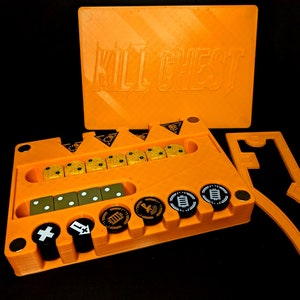 May include: An orange board game organizer with the text "KILL CHEST" on the lid. The organizer holds dice, tokens, and other game components. The game pieces are yellow, black, and white.