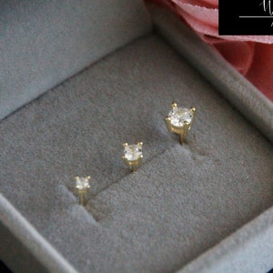 May include: Three gold-toned earrings with clear gemstones in a gray velvet jewelry box. The box has the text "Whispers and Bijoux" on the top.