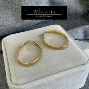 May include: A pair of gold oval hoop earrings. The earrings are presented on a grey velvet surface. The brand name "Whispers and Bijoux" is displayed in a black rectangle.