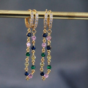Könnte beinhalten: Goldfarbene Creolen mit einer herabhängenden Kette aus bunten Edelsteinen. Die Reifen sind mit kleinen, klaren Kristallen besetzt. Die Edelsteine sind in den Farbtönen Blau, Rosa, Grün und Gelb gehalten.