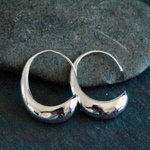 May include: A pair of polished silver hoop earrings with a crescent shape. The earrings are displayed on a dark gray stone surface, reflecting light. The design is minimalist and modern, suitable for everyday wear.
