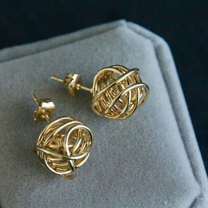 May include: A pair of gold-toned earrings with a knotted sphere design. The earrings are displayed on a grey velvet surface.