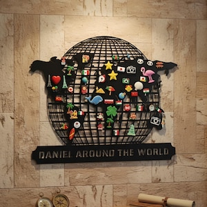 Peut inclure: Décoration murale en métal noir représentant une carte du monde en forme de globe, ornée de divers aimants colorés sur le thème du voyage. Le texte "DANIEL AROUND THE WORLD" est affiché en dessous. Sur un mur de briques brun clair.