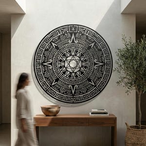 Può includere: Una grande opera d'arte murale circolare in metallo nero con un intricato design geometrico, simile a un sole o a un calendario. L'opera d'arte è montata su una parete bianca, sopra una consolle in legno. Una donna passa accanto all'opera.