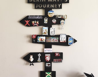 May include: Black wooden signpost with the text "DERIN MAVI'S JOURNEY" and arrows pointing in different directions. The arrows are decorated with travel-themed magnets from various locations, including Amsterdam, London, and San Francisco.