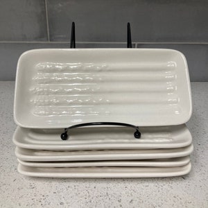 May include: A set of six white ceramic rectangular serving dishes with a textured surface. The dishes are stacked on top of each other and are held together by a black metal stand.