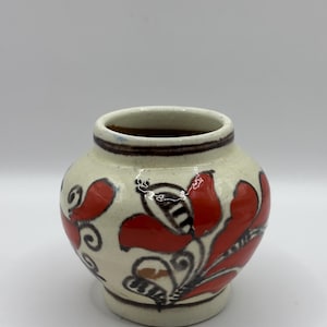 May include: A small, round ceramic vase with a cream-colored base. The vase is adorned with red floral designs and black accents. The rim features a brown band. This decorative vase is suitable for small flowers or as a decorative accent.