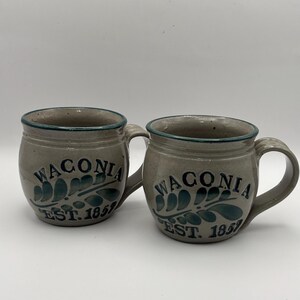 May include: Two gray ceramic mugs with green and blue designs. The mugs have the text "Waconai Est. 1857" on them.