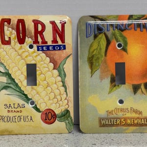 May include: Two vintage-style light switch covers. One features a corn cob with the text "CORN SEEDS" and "SALAS BRAND PRODUCE OF U.S.A." The other depicts oranges with the text "DISTINCTIVE BRAND" and "THE CITRUS FARM OF WALTER S. NEWHALL."