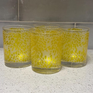 May include: Four clear drinking glasses with a yellow floral pattern. The glasses are cylindrical and sit on a white surface. The background is a light gray tiled wall.