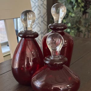 May include: Three decorative glass bottles in varying sizes. The bottles are a deep red color with clear glass stoppers. The bottles are arranged on a dark wooden surface, suggesting a home decor theme.