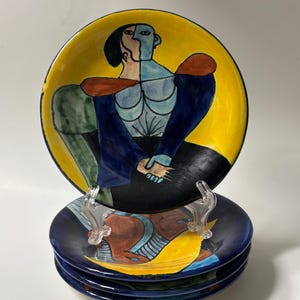May include: A set of decorative ceramic plates with a unique abstract design. The plates feature a yellow background with a figure in blue, black, and red. The set includes four plates, stacked on top of each other.