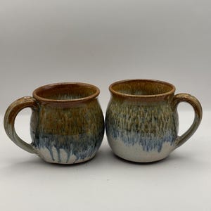 May include: Two handmade ceramic mugs with a brown, blue, and white glaze. The mugs have a rounded body and a handle.
