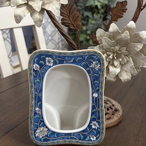 May include: A rectangular ceramic picture frame with a blue floral design and gold trim. The frame has a white interior and is set on a wooden surface. Decorative floral accents are in the background.