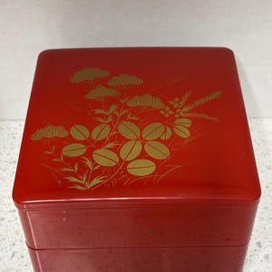 May include: A red, square, two-tiered box with a glossy finish. The lid is decorated with a gold floral design. The box has a simple, elegant design, suitable for storage or display.