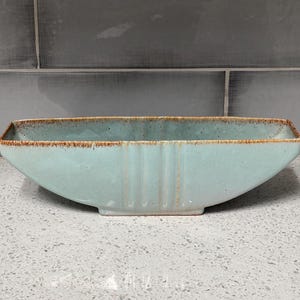 May include: A rectangular, light blue ceramic bowl with a slightly curved shape. The rim has a brown, textured edge. Vertical lines are etched into the front of the bowl. The bowl is sitting on a speckled, light gray surface.