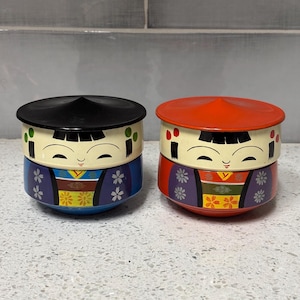 May include: Two decorative Japanese Kokeshi doll-shaped containers. One has a black lid and blue base, the other has a red lid and orange base. Both have painted faces and floral patterns.