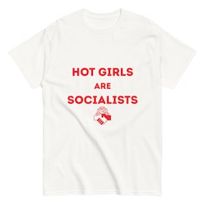 Puede incluir: Una camiseta blanca de manga corta con el texto rojo "HOT GIRLS ARE SOCIALISTS" impreso en el pecho. Debajo del texto hay un icono gráfico rojo que representa una rosa situada sobre dos manos entrelazadas en un apretón de manos.
