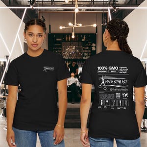 May include: Black t-shirts with white text and graphics. The front of the shirt has the words "HAIR STYLIST" and a scissors graphic. The back of the shirt has the words "100% GMO FREE ORGANIC" and more text and graphics.
