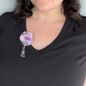 May include: A round badge reel with the words "Speech Therapy" in purple script. The badge reel has a floral design with pink, purple, and blue flowers. The reel is attached to a black shirt.