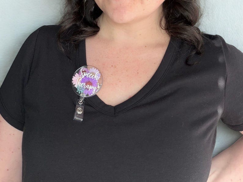 May include: A round badge reel with the words "Speech Therapy" in purple script. The badge reel has a floral design with pink, purple, and blue flowers. The reel is attached to a black shirt.