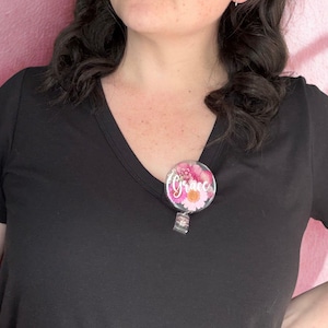 May include: A round, floral-themed badge reel with the name "Grace" in white script. The badge features pink and white flowers. The reel is attached to a black V-neck shirt.