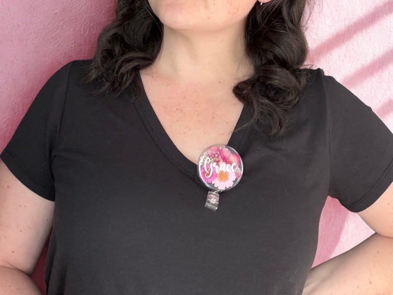 May include: A round, floral-themed badge reel with the name "Grace" in white script. The badge features pink and white flowers. The reel is attached to a black V-neck shirt.