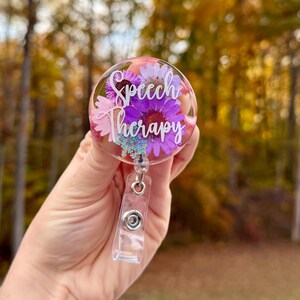 May include: A clear badge reel with the words "Speech Therapy" in white script. The badge reel is filled with purple and pink flowers and small blue accents. The reel is held in a hand against a blurred background of fall foliage.