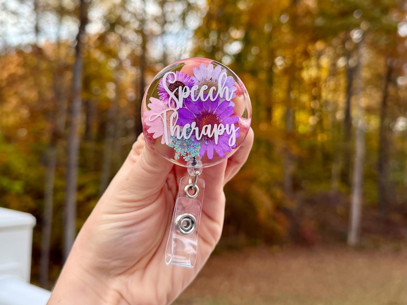 May include: A clear badge reel with the words "Speech Therapy" in white script. The badge reel is filled with purple and pink flowers and small blue accents. The reel is held in a hand against a blurred background of fall foliage.