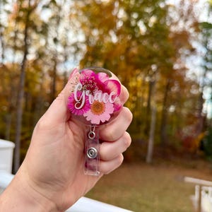 May include: A clear, round badge reel with a floral design featuring pink and white flowers. The name "Grace" is written in cursive across the front. The reel has a retractable cord and a metal clip.