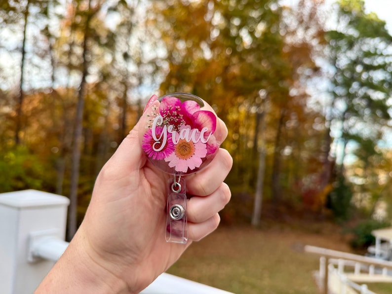 May include: A clear, round badge reel with a floral design featuring pink and white flowers. The name "Grace" is written in cursive across the front. The reel has a retractable cord and a metal clip.