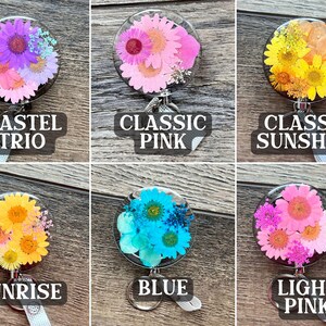 May include: A collection of six floral badge reels with clear resin domes. Each reel features a different arrangement of dried flowers in various colors, including pink, yellow, and blue. Each reel has a label with a unique name: Pastel Trio, Classic Pink, Classic Sunshine, Sunrise, Blue, and Light Pink.