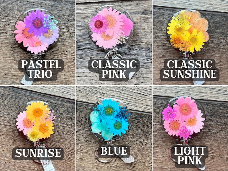 May include: A collection of six floral badge reels with clear resin domes. Each reel features a different arrangement of dried flowers in various colors, including pink, yellow, and blue. Each reel has a label with a unique name: Pastel Trio, Classic Pink, Classic Sunshine, Sunrise, Blue, and Light Pink.