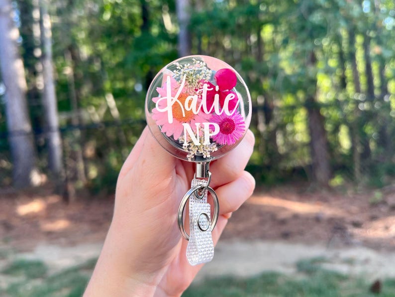 May include: A clear, round badge reel with a floral design and the name "Katie NP" in white script. The reel features a retractable cord and a metal clip for attachment. The background is a blurred view of green trees.