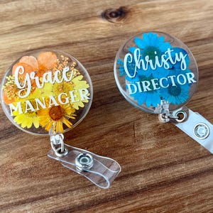 May include: Two clear acrylic badge reels with floral designs. One features orange and yellow flowers with the text "Grace MANAGER". The other has blue flowers and the text "Christy DIRECTOR". Both have clear plastic straps.