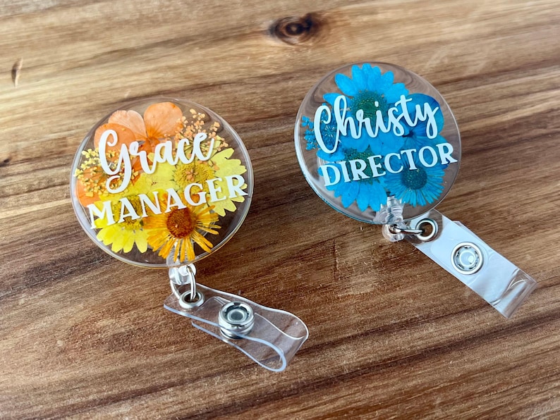 May include: Two clear acrylic badge reels with floral designs. One features orange and yellow flowers with the text "Grace MANAGER". The other has blue flowers and the text "Christy DIRECTOR". Both have clear plastic straps.