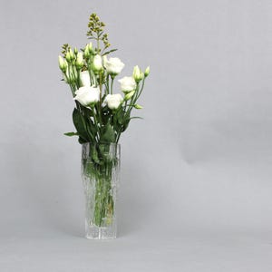 Könnte beinhalten: Eine klare Glasvase mit strukturierter Oberfläche hält einen Strauß weißer Blumen mit grünen Stielen. Die Vase steht auf einem grauen Hintergrund.