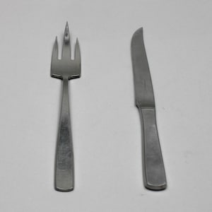 Könnte beinhalten: Ein Edelstahl-Gabel- und Messerset. Die Gabel hat drei Zinken und einen rechteckigen Griff. Das Messer hat eine gebogene Klinge und einen passenden Griff. Beide Teile sind aus gebürstetem Metall gefertigt.