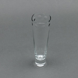 Puede incluir: Un vaso transparente y delgado con una parte superior acampanada y una base redondeada. El borde superior presenta tres pequeñas hendiduras circulares. El vaso tiene un aspecto liso y transparente, adecuado para servir bebidas. El vaso mide unos 18 cm de alto.