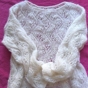 May include: Ivory-colored, open-weave sweater with long sleeves. The knitwear features a delicate, lacy pattern across the bodice and sleeves. The sweater is displayed on a bright pink surface, showcasing its intricate design.