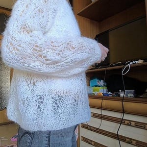 May include: A white, open-weave sweater with long sleeves. The knitwear has a delicate, lacy pattern. The sweater is worn over a pair of blue jeans. The image is taken indoors, with a wooden door and cabinet in the background.