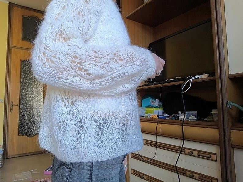 May include: A white, open-weave sweater with long sleeves. The knitwear has a delicate, lacy pattern. The sweater is worn over a pair of blue jeans. The image is taken indoors, with a wooden door and cabinet in the background.