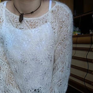 May include: A cream-colored, open-weave sweater with a round neckline. The sweater has a delicate, lacy pattern and long sleeves. A dark brown pendant hangs from a black cord necklace. The sweater is worn over a white top.