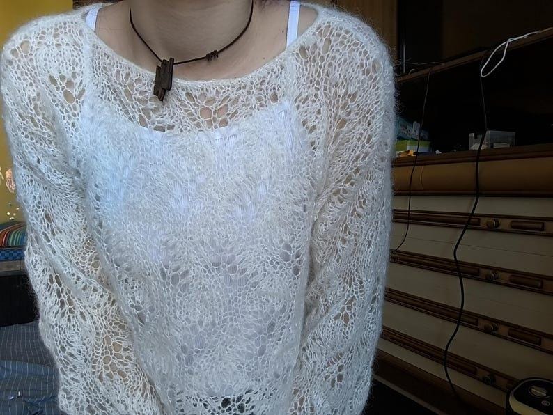 May include: A cream-colored, open-weave sweater with a round neckline. The sweater has a delicate, lacy pattern and long sleeves. A dark brown pendant hangs from a black cord necklace. The sweater is worn over a white top.