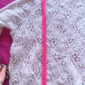 May include: A white, open-weave sweater with long sleeves. The sweater's length is measured with a pink measuring tape, showing a length of approximately 51 inches. The knit pattern features a delicate, lacy design.