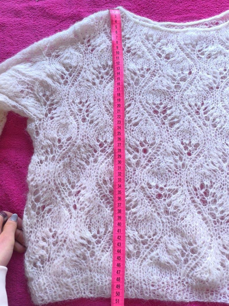 May include: A white, open-weave sweater with long sleeves. The sweater's length is measured with a pink measuring tape, showing a length of approximately 51 inches. The knit pattern features a delicate, lacy design.