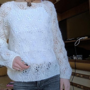 May include: A white, open-weave sweater with long sleeves. The sweater has a delicate, lacy pattern throughout, creating a semi-sheer effect. The neckline is a simple scoop, and the overall style is casual and comfortable. The sweater is paired with blue jeans.