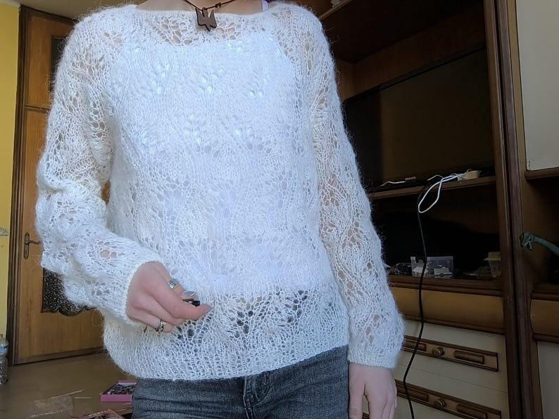 May include: A white, open-weave sweater with long sleeves. The sweater has a delicate, lacy pattern throughout, creating a semi-sheer effect. The neckline is a simple scoop, and the overall style is casual and comfortable. The sweater is paired with blue jeans.