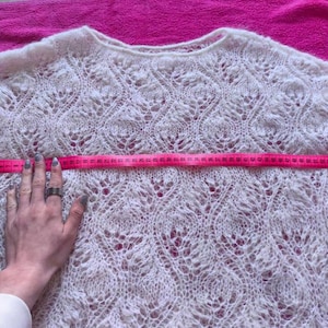 May include: A cream-colored, open-weave sweater with a round neckline. A pink measuring tape is stretched across the chest of the sweater. The sweater's width is being measured. The sweater has a delicate, lacy pattern.